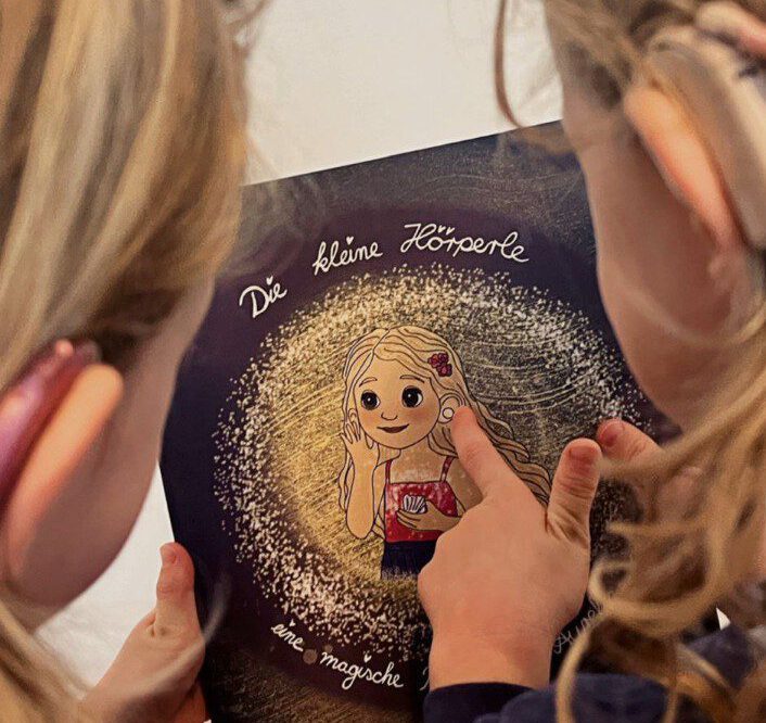 Zwei Kinder mit Hörhilfen schauen gemeinsam in das Kinderbuch Die kleine Hörperle.